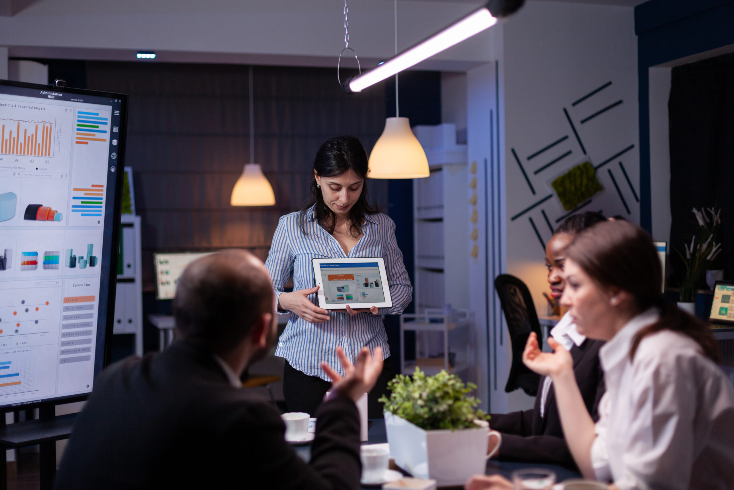 Overworked workaholic entrepreneur woman shwoing marketing graphs using tablet overworking at company solution late at night in meeting room. Diverse multi-ethnic teamwork solving financial strategy